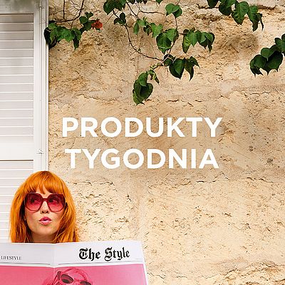 Kobieta z rudymi włosami i różowymi okularami przeciwsłonecznymi czyta gazetę o modzie „The Style” na tle kamiennej ściany z napisem „PRODUKTY TYGODNIA”.