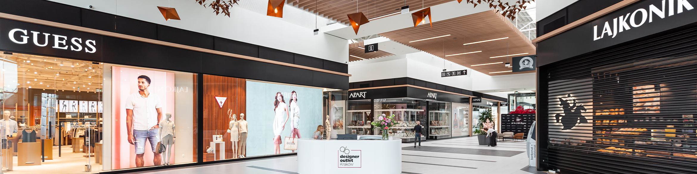 Nowoczesne wnętrze centrum handlowego ze sklepami GUESS, Apart i Lajkonik, z eleganckim wystrojem i centralnym punktem informacyjnym.
