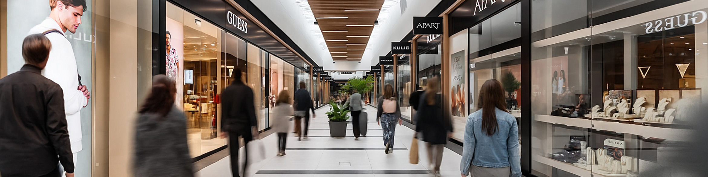 Wnętrze centrum handlowego Designer Outlet Kraków z butikami znanych marek po obu stronach, takimi jak GUESS, APART i KULIG, oraz spacerującymi klientami w nowoczesnej, jasnej przestrzeni.
