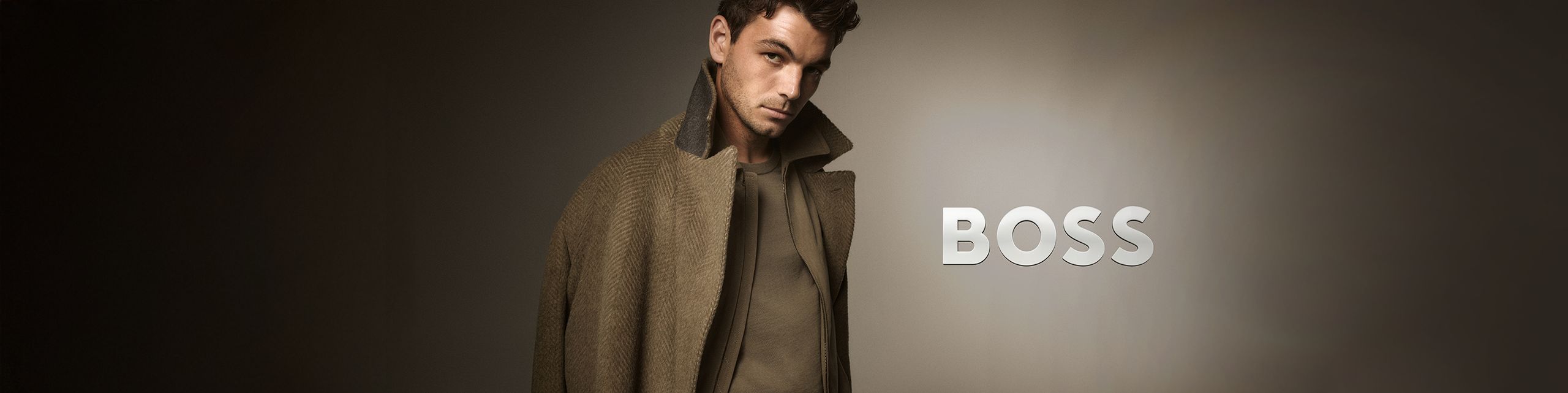 Mężczyzna w długim, eleganckim płaszczu w odcieniach brązu patrzy w stronę aparatu; po prawej stronie znajduje się logo ‘BOSS’.