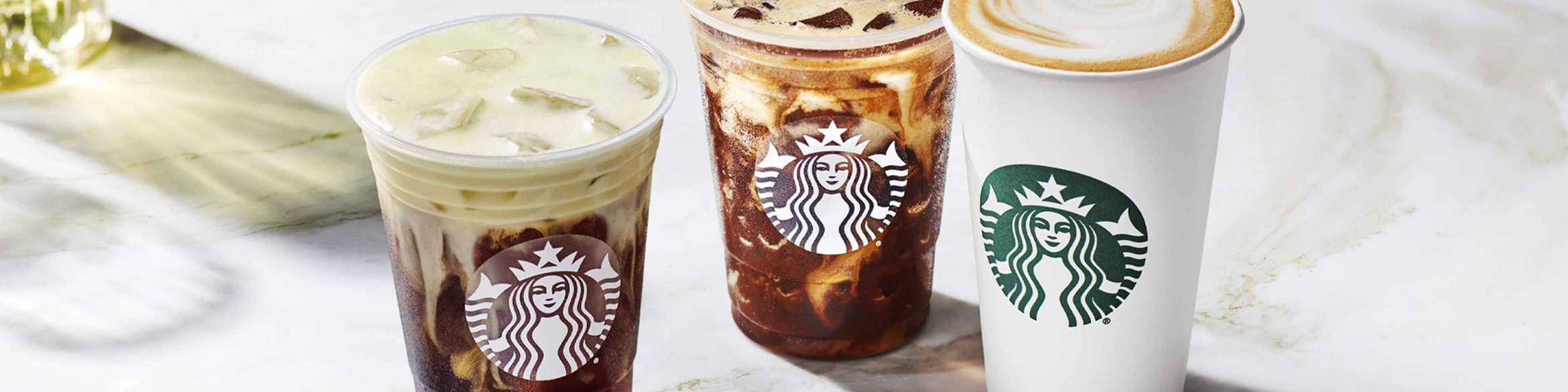 Trzy napoje z logo Starbucks ustawione na marmurowym blacie – dwa w plastikowych kubkach z lodem (jeden jasny, drugi ciemny) oraz jeden w białym papierowym kubku z mleczną pianką.